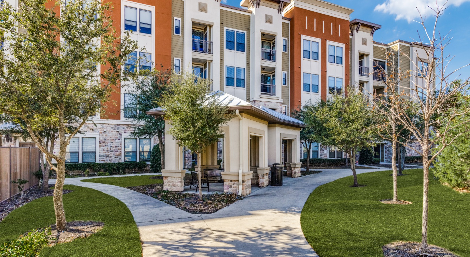 the front of an apartment complex with landscaping and trees at The  Carson @ Twin Creeks